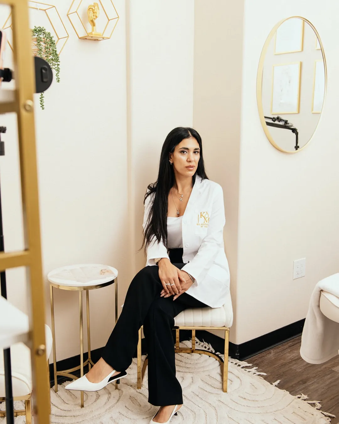Koral, founder of KM RX Aesthetics, seated in the Bellaire studio beside the gold mirror